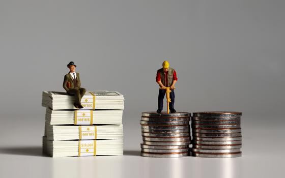 Figurine of a businessman sits on a pile of $100 bills while the figurine of a construction worker works on a shorter pile of coins (Dreamstime/Hye Jin Kang)