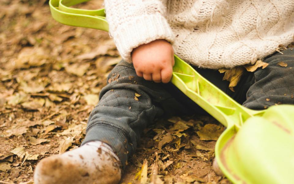 A cropped view of a baby wearing a white sweater and grey pants covered in dirt while holding a green plastic shovel and sitting in a pile of brown leaves.